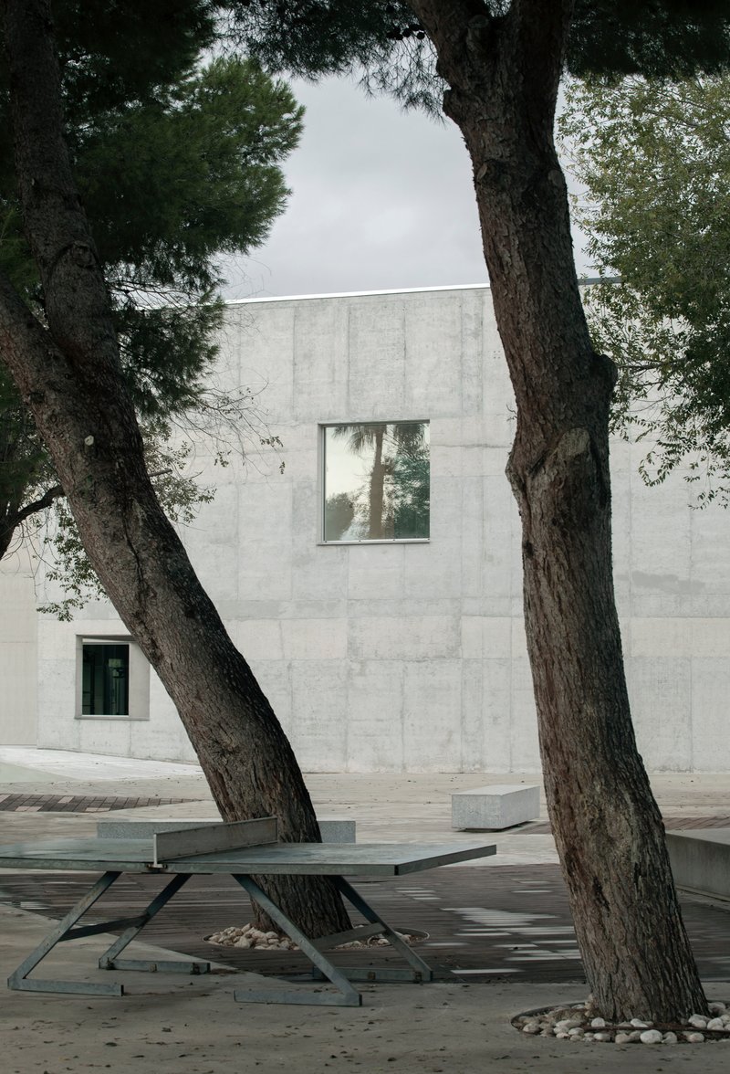 Metal picnic table beneath pine trees facing a board-formed concrete facade with square window openings