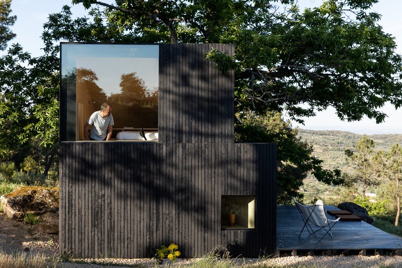 Charred timber volume with large window framing an occupant, nestled among oak trees on a hillside