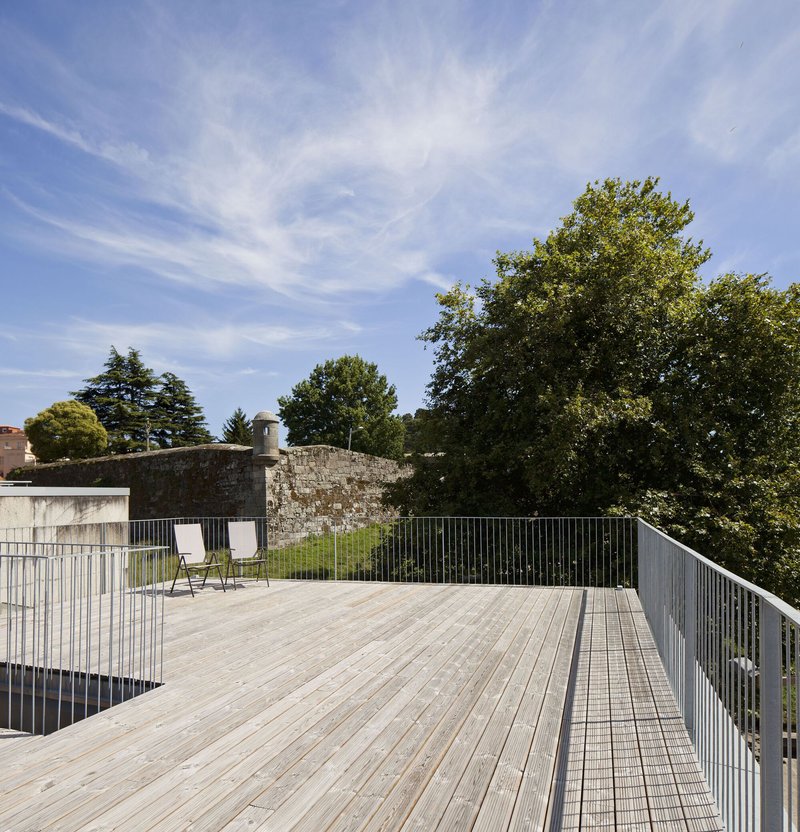 Timber deck terrace with metal mesh railings beside a stone wall and mature trees under open sky