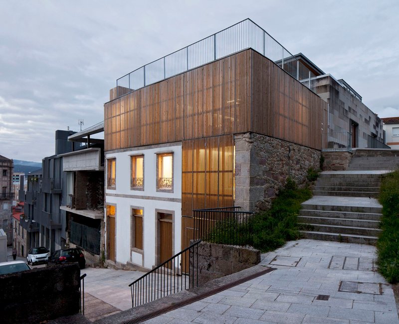 Corner facade showing timber and corrugated metal volumes rising above a stone retaining wall at twilight