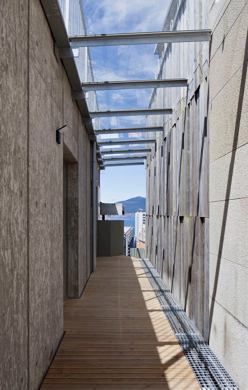 Timber deck corridor with overhead glass canopy between wood-clad and concrete walls framing a harbor view