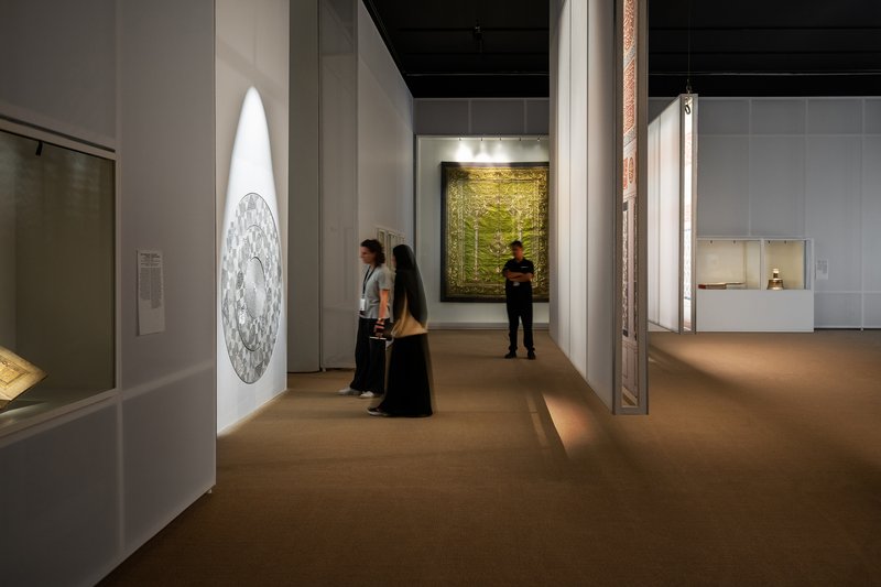 Gallery corridor with visitors viewing illuminated artworks on white walls and glass display cases