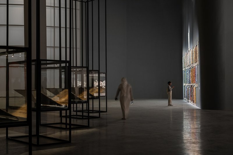 Gallery hall with black steel frame shelving and visitors near illuminated textile hanging on wall