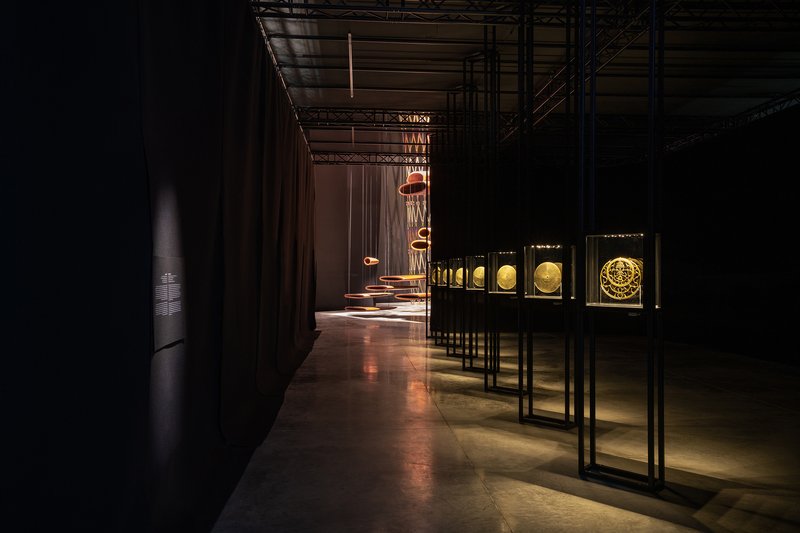Exhibition corridor with backlit artifact cases and suspended copper lighting elements under exposed ceiling infrastructure