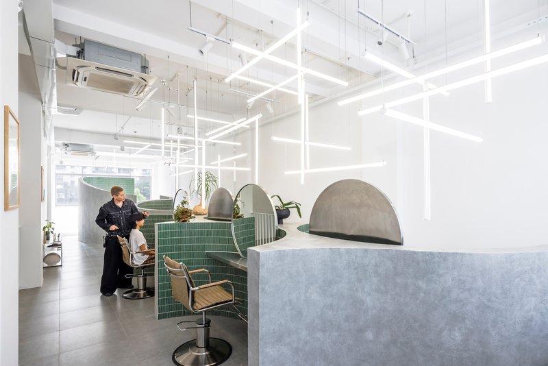 White grid light fixture suspended over green tiled salon stations with chrome chairs and a stylist at work