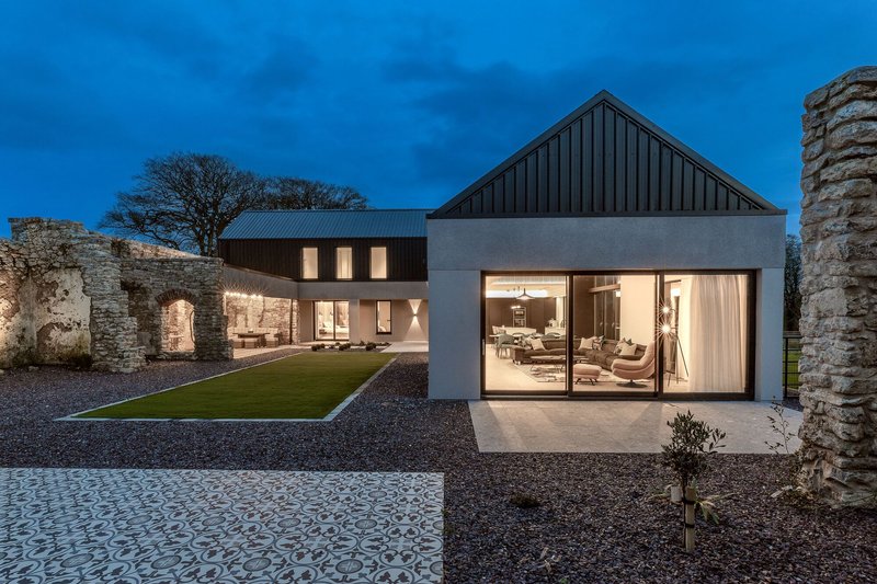 Glass living pavilion opening to the courtyard with metal roofed volumes and stone walls at dusk