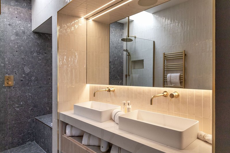 Bathroom vanity with dual vessel sinks, backlit vertical tiles, and brass fixtures beneath a timber-lined ceiling