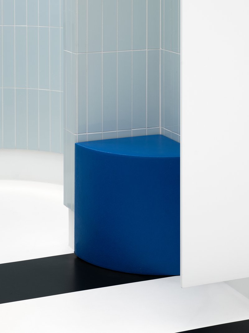 Blue upholstered corner bench against pale blue tiled walls and black striped floor