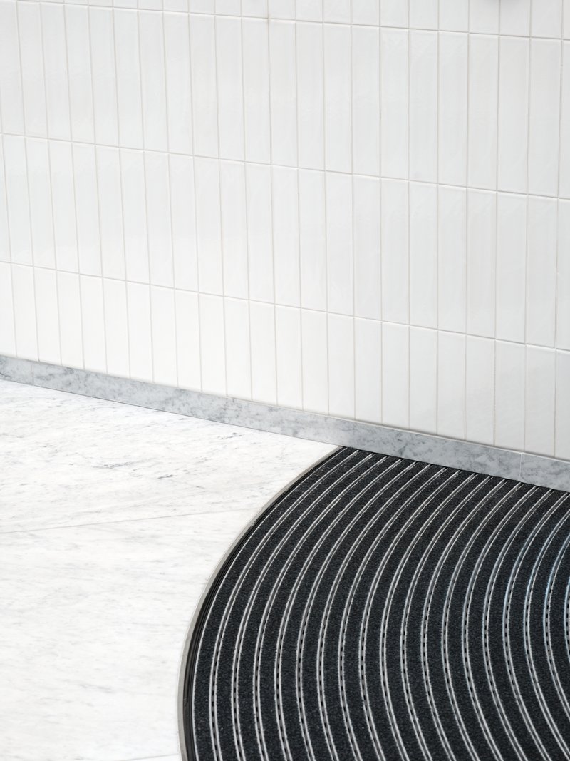 Curved black ribbed floor mat meeting white vertical tile wall with terrazzo base trim
