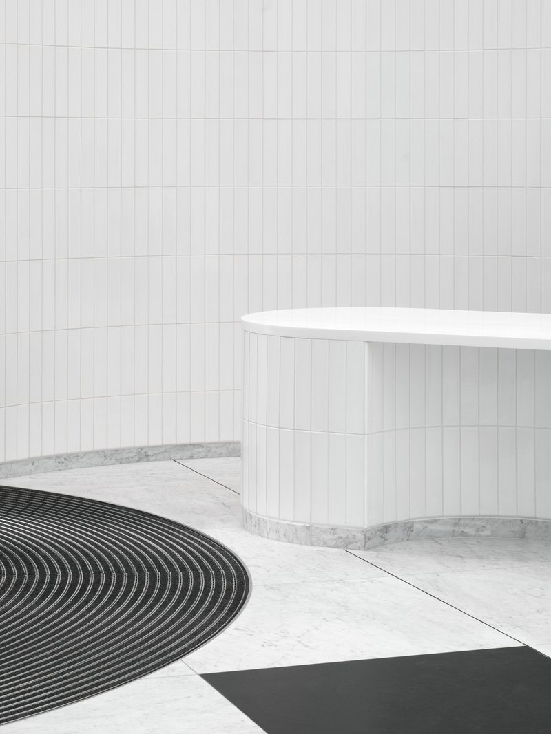 White cylindrical reception desk beside curved black floor mat on terrazzo in a tiled lobby