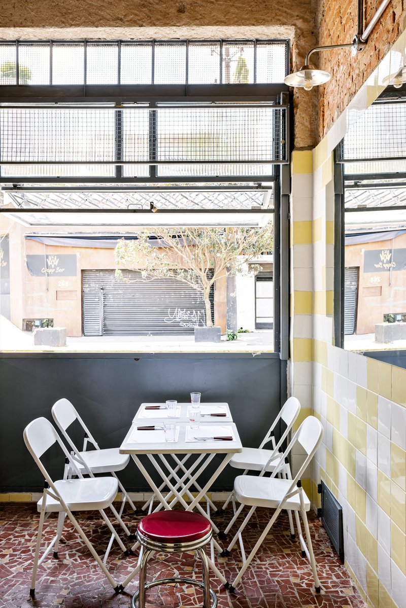 Dining table with white folding chairs positioned below a steel-framed window overlooking the street