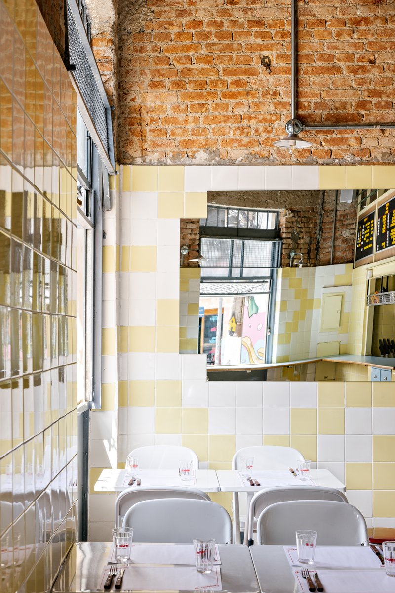 Dining tables beneath exposed brick and a mirrored wall reflecting yellow and white checkerboard tile surfaces