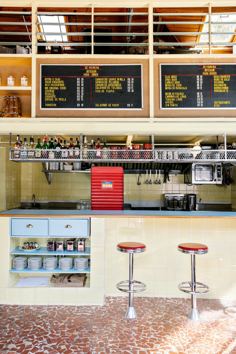 Interior bar counter with yellow tile wainscoting, chrome stools, digital menu boards and perforated metal shelf