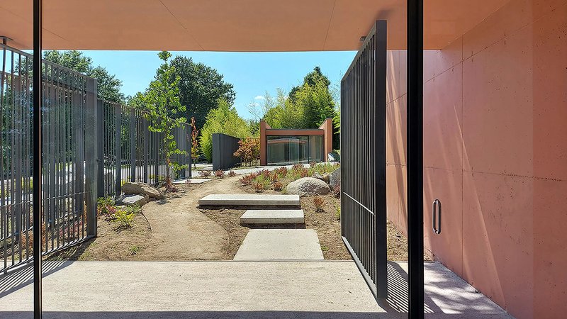 View through covered passage showing metal gate, courtyard steps, and planted beds with distant pavilion under blue sky