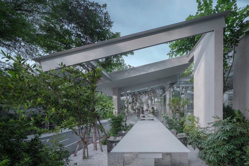 Elevated concrete walkway flanked by planted beds and trees under intersecting white beam structure