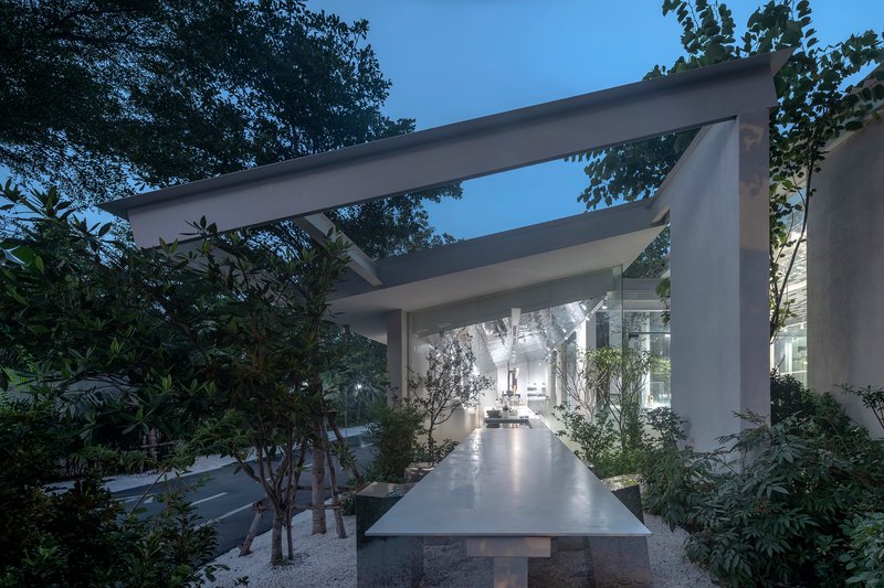 Long concrete table beneath horizontal beam structure surrounded by planted beds at dusk