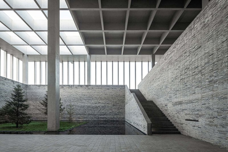 Interior courtyard with stone walls, concrete columns supporting a coffered skylight, and a staircase under diffused light