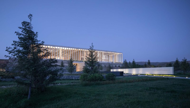 Slatted upper volume set on a masonry base surrounded by lawn and conifer trees at twilight