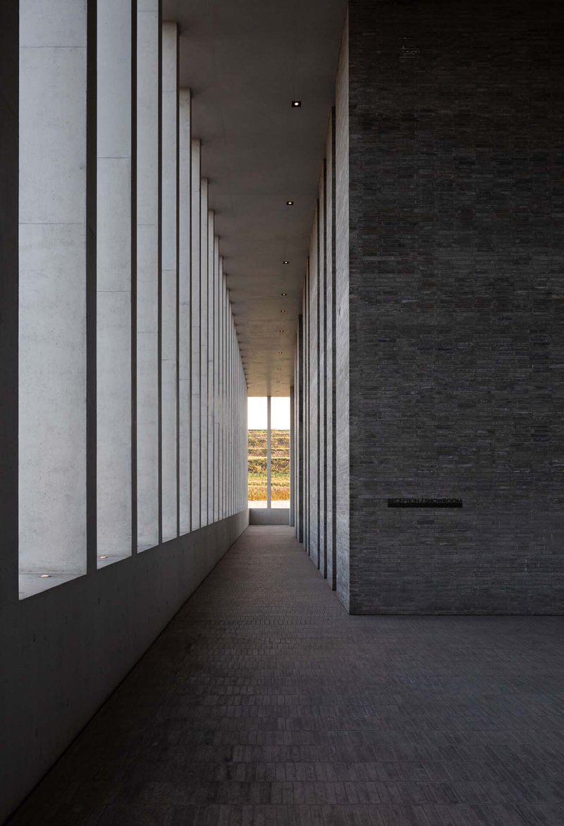 Long corridor with rhythmic vertical concrete fins casting shadows on dark brick flooring at dusk