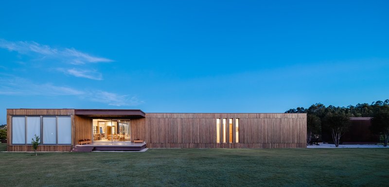 Horizontal timber-clad volume with vertical window slits across an open lawn at dusk