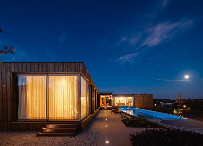 Glazed bedroom volume with curtains beside a pool and planted beds at dusk