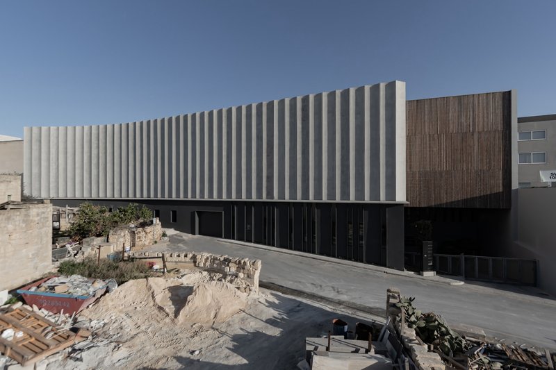 Facade with white vertical louvres and timber-clad volume rising above a paved forecourt under clear sky