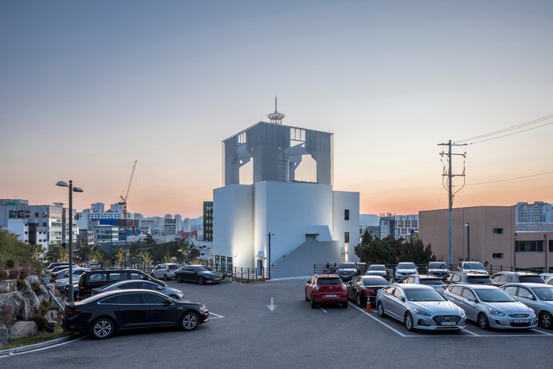 White tower with arched openings illuminated at dusk beside a parking lot and utility poles