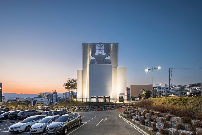 Illuminated cylindrical tower housing a bell in front of a parking area at sunset