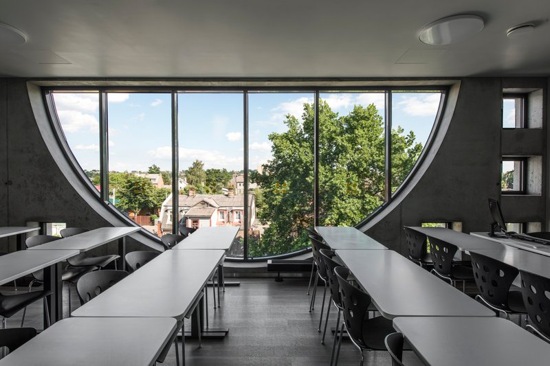Interior classroom with rows of tables facing a semicircular window overlooking leafy trees and rooftops