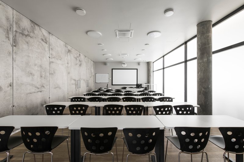 Classroom with rows of white tables and black perforated chairs facing projection screen