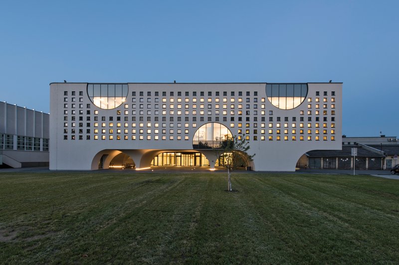 Front elevation showing the gridded white facade with illuminated arched entrance portals at dusk