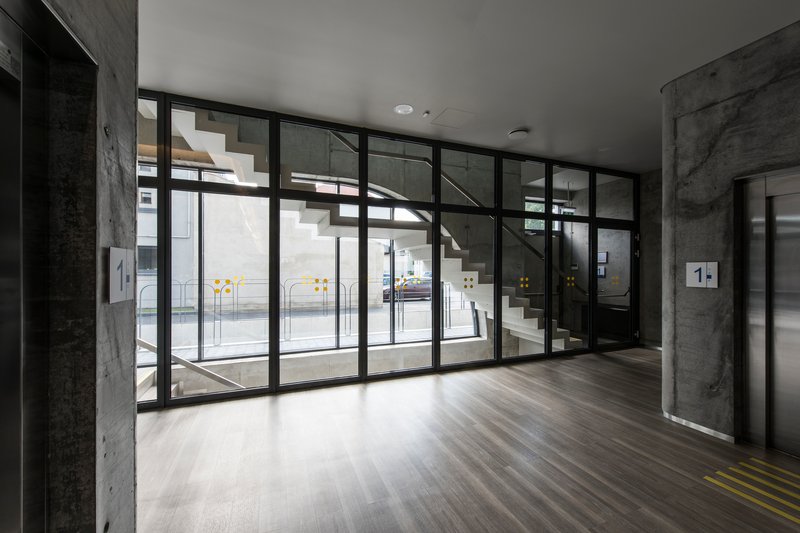 Lobby entrance with black-framed glazing overlooking white staircase and exposed concrete columns