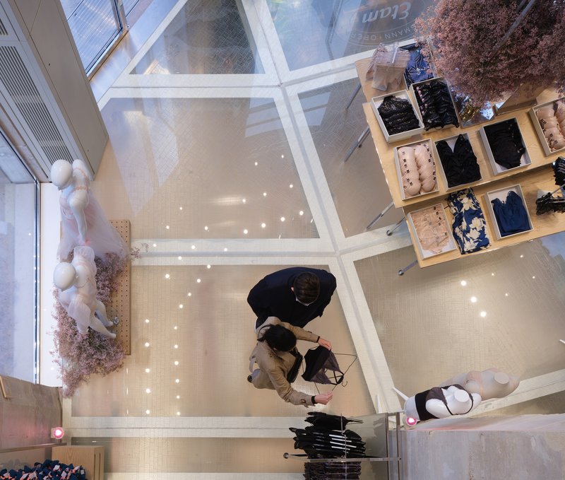 Top-down view through glass atrium ceiling showing shoppers on the terrazzo floor with merchandise displays