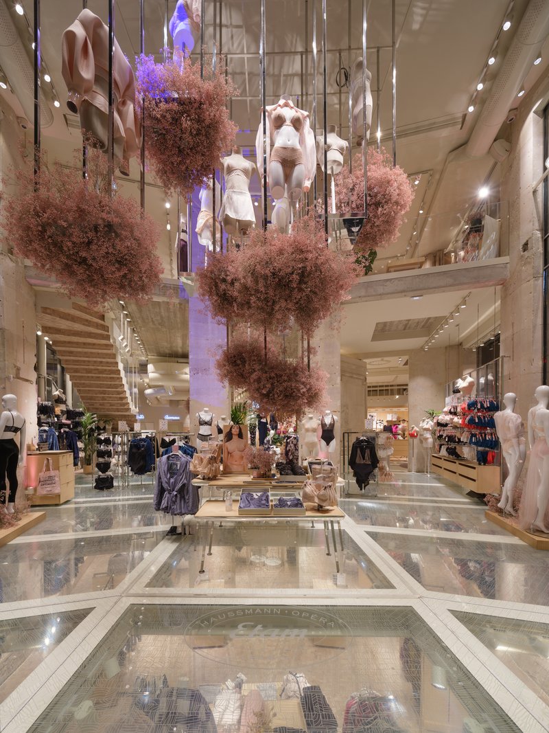 Double-height retail atrium with suspended pink foliage and glass floor panels revealing the level below