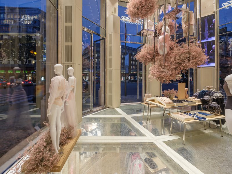 Street-level storefront at dusk with floor-to-ceiling glazing and suspended pink foliage inside