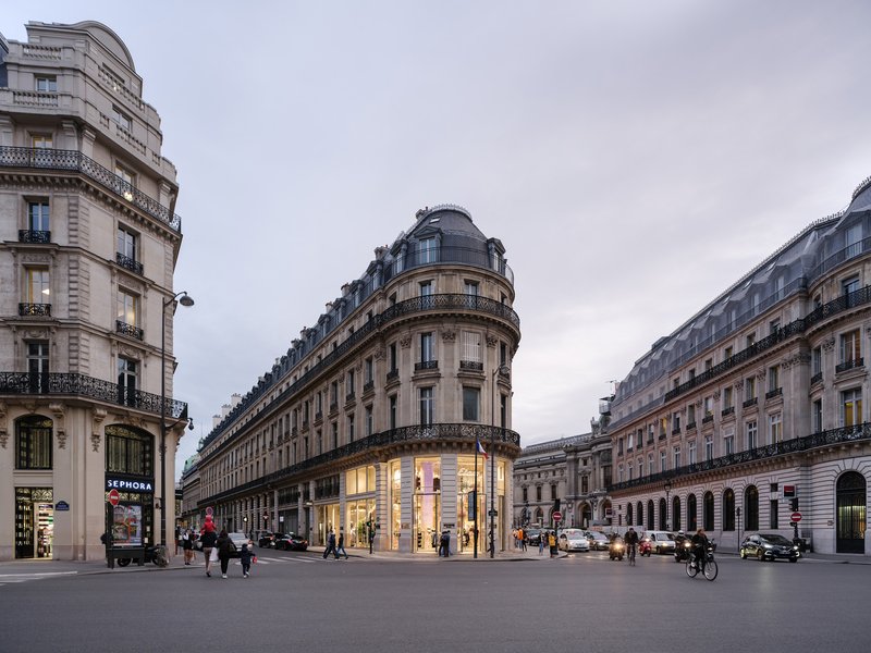 Corner retail building with illuminated ground floor windows at dusk surrounded by pedestrians and cyclists