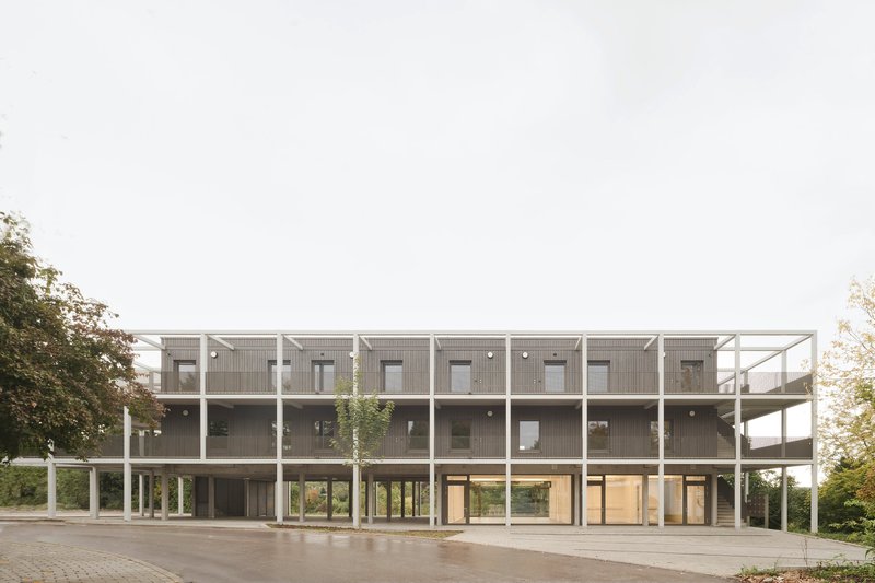 Front elevation showing the repeating module facade with white structural frame and concrete columns on a paved plaza