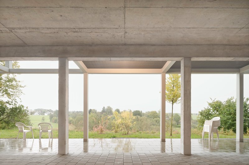 Covered ground-level terrace with exposed concrete ceiling and white plastic chairs overlooking fields and trees