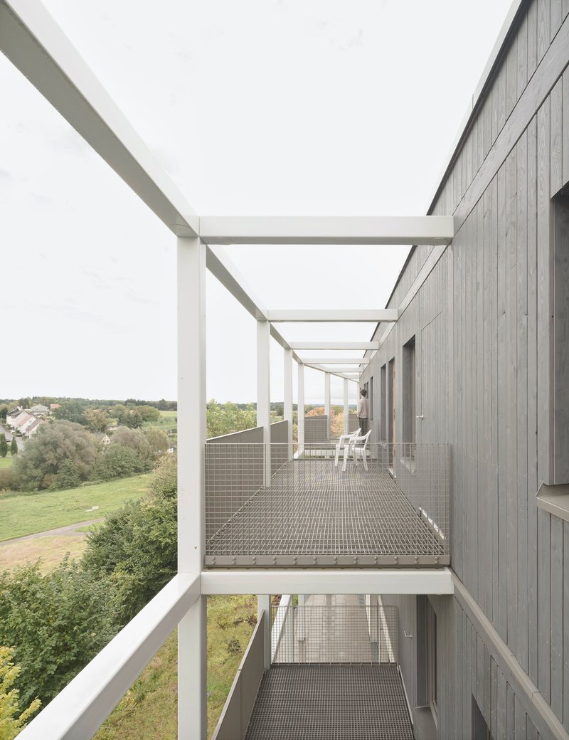 Exterior walkway with steel grating deck and white frame pergola overlooking a landscape of fields and trees
