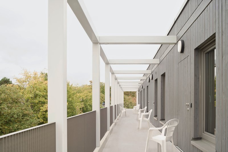 Exterior walkway with white steel pergola, grey cladding and white plastic chairs overlooking green trees