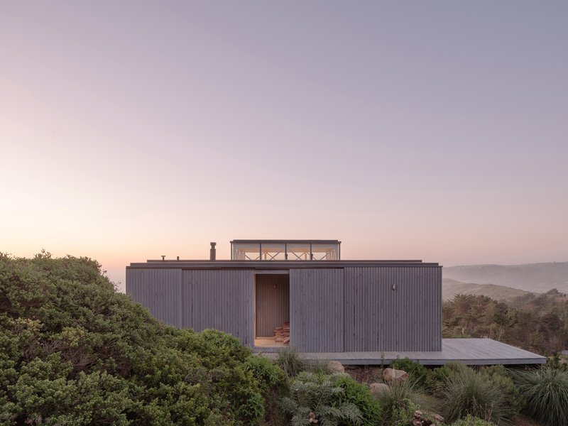 Vertically ribbed timber-clad volume with rooftop terrace overlooking coastal hills at dusk