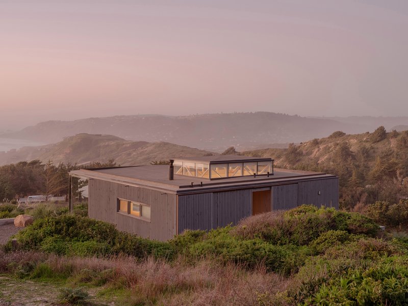Corrugated metal residence with glazed rooftop volume nestled into hillside scrub at sunset