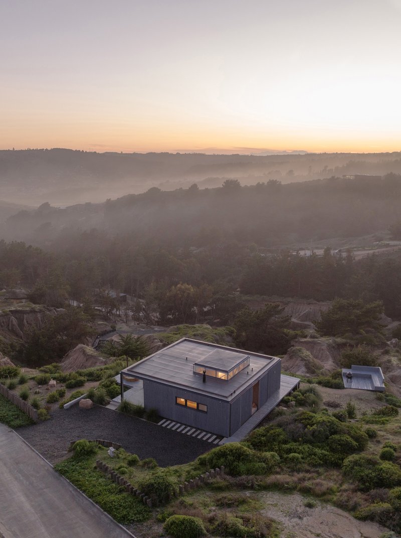 Aerial view of the corrugated metal-clad volume nestled among rocky hillside vegetation at dusk with mist