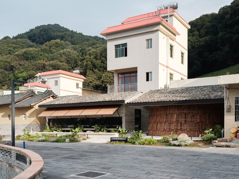 Low-slung pavilion with timber-shingled roof and planted courtyard beside paved plaza and hillside housing