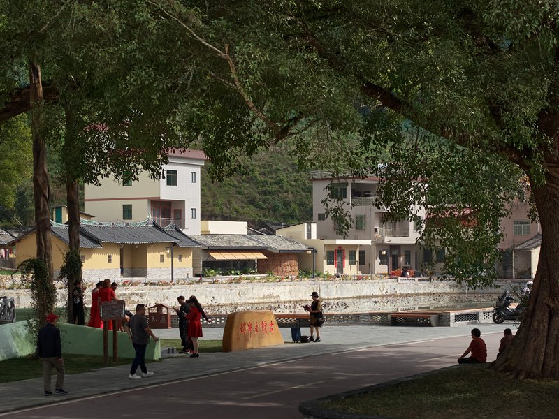 Low tiled roof pavilion viewed from plaza with visitors gathered beneath mature trees