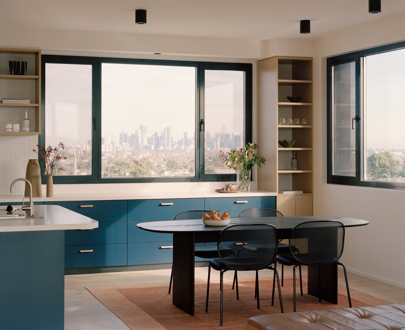 Open-plan kitchen and dining area with blue cabinetry and black-framed windows overlooking the city skyline