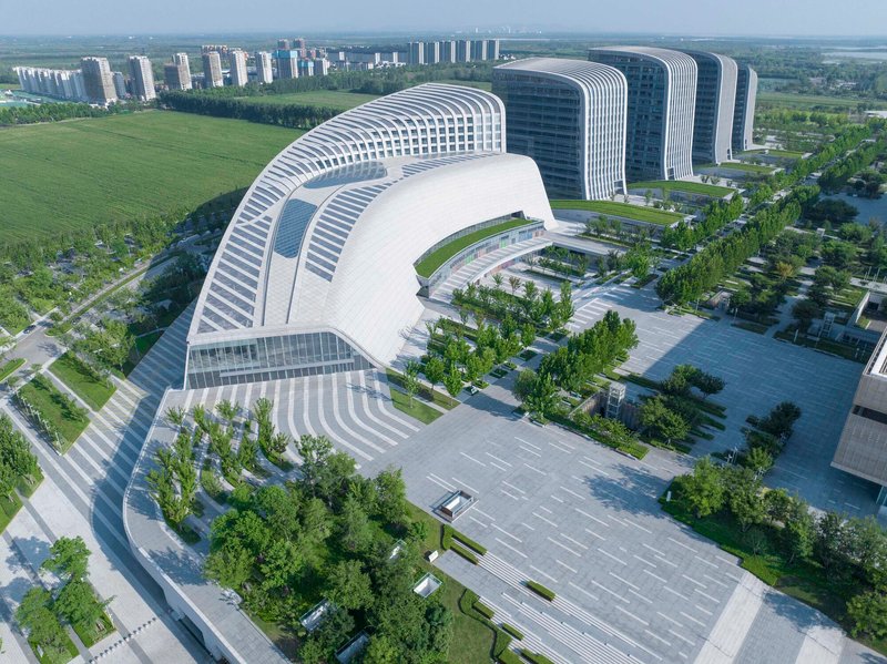 Curved building with horizontal white louvers wrapping a courtyard and entrance plaza under a clear blue sky