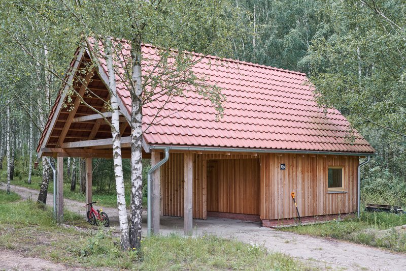Timber cabin with red tile roof and open carport framed by birch trees on a gravel clearing