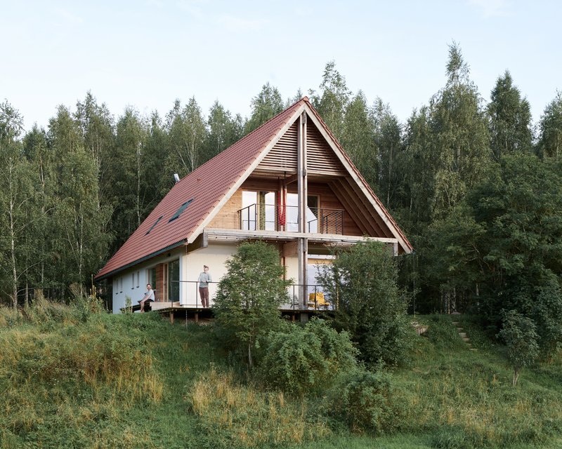 Steeply pitched red metal roof with wood cladding and balcony visible against a dense tree backdrop