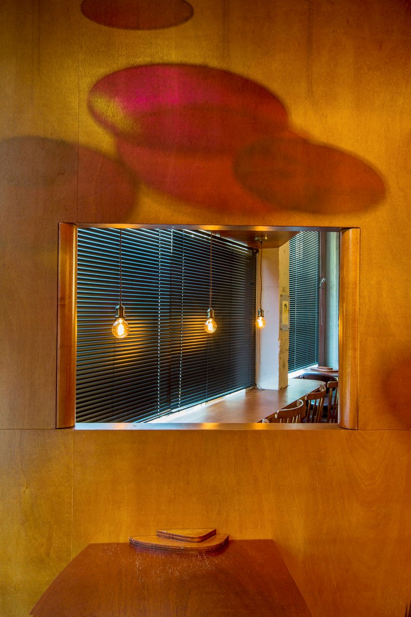 Framed aperture in wood-paneled wall revealing slatted metal screens and pendant lights with reflected colored light shadows